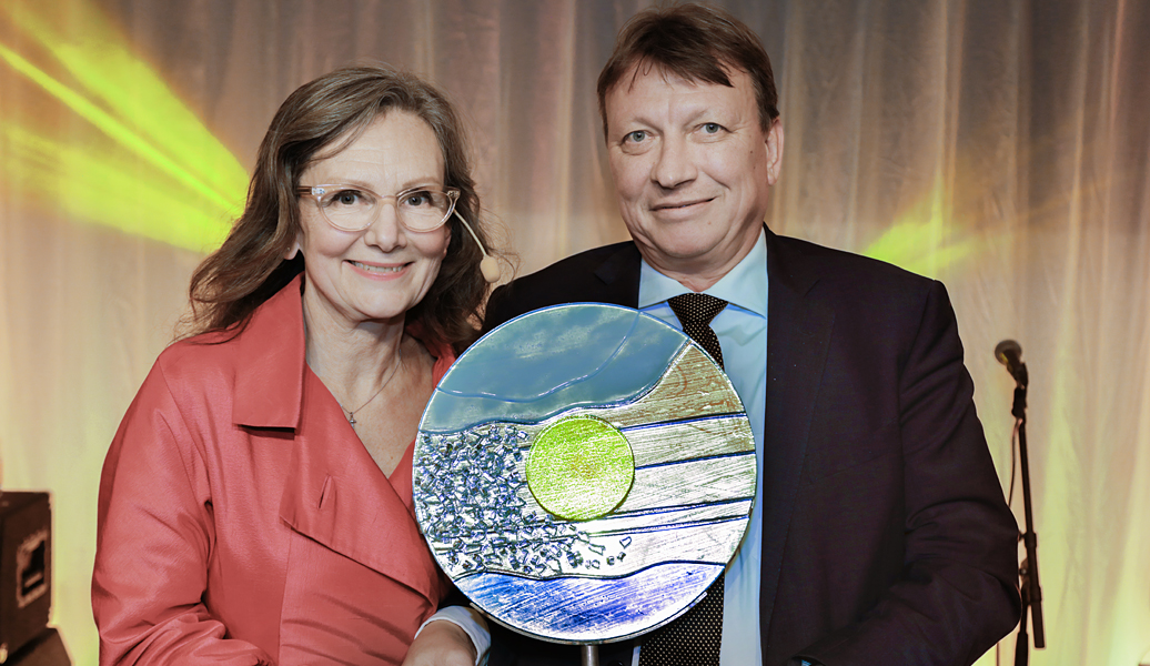 En kvinne og en mann smiler til kamera og holder en pris i resirkulert glass.